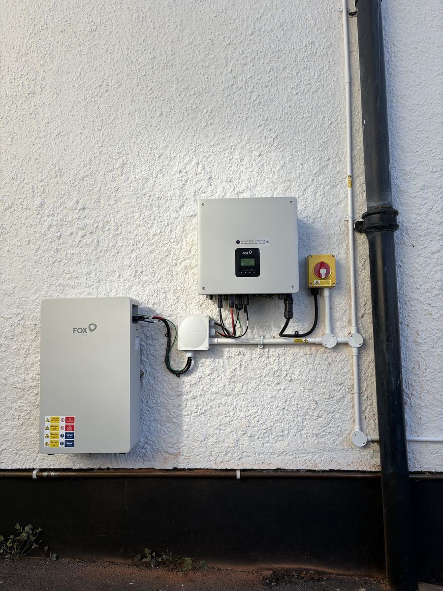 Fox ESS hybrid inverter and EP6 Plus battery mounted on a white rendered outside wall, with an isolator and cabling in white conduit